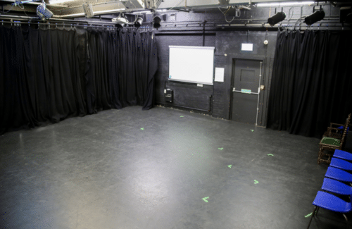 Image shows a drama studio for hire in Harrow. A largely black room with black walls and floors adorned with black equipment. Also in view are a line of blue chairs and profesionally rigged lighting equipment on bars suspended from the ceiling.